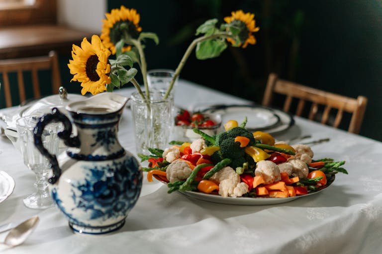 A beautifully set dining table with fresh vegetables, sunflowers, and an ornate jug creates a cozy atmosphere.
