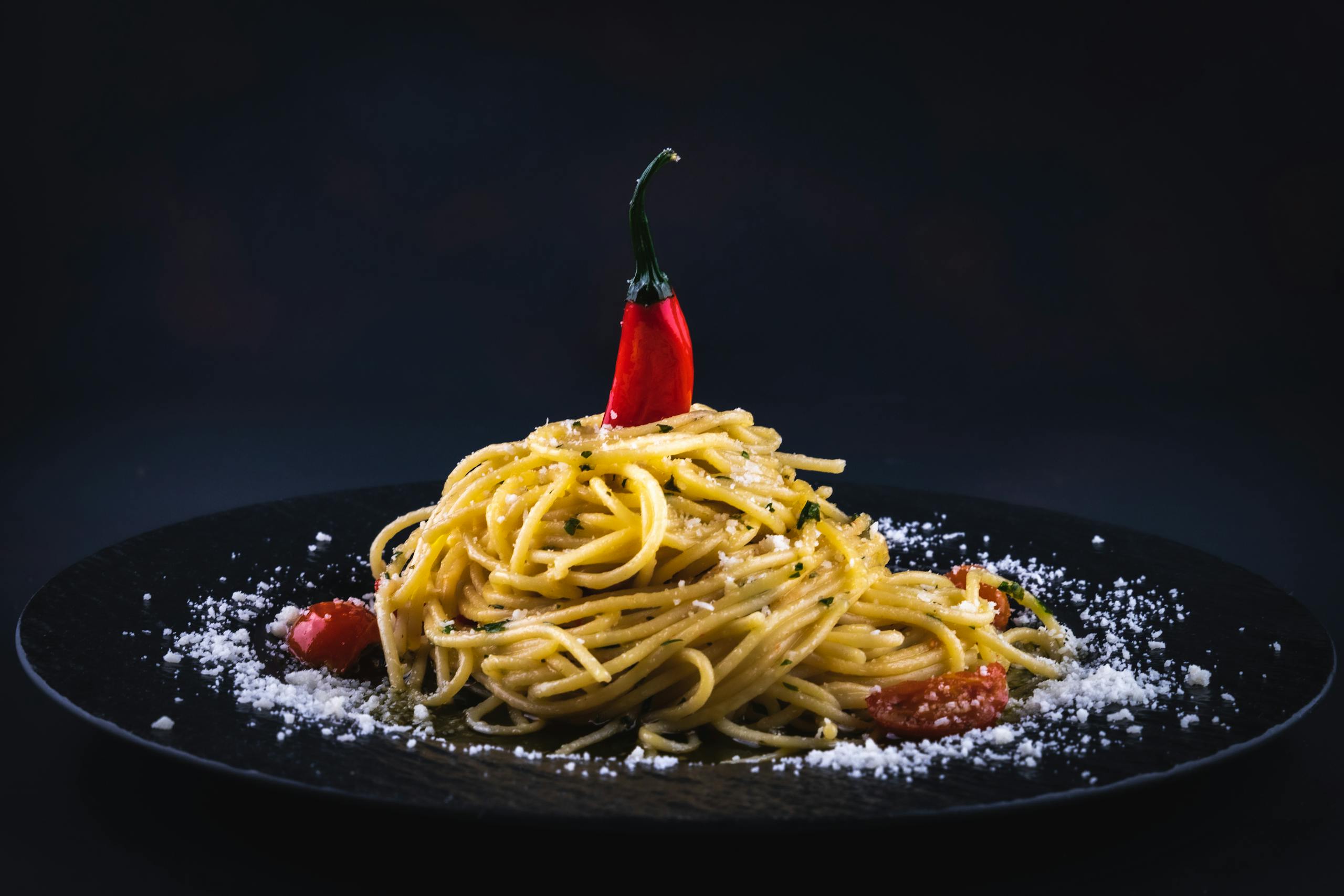 A beautifully styled plate of spaghetti with chili pepper on a black dish, garnished with cheese.