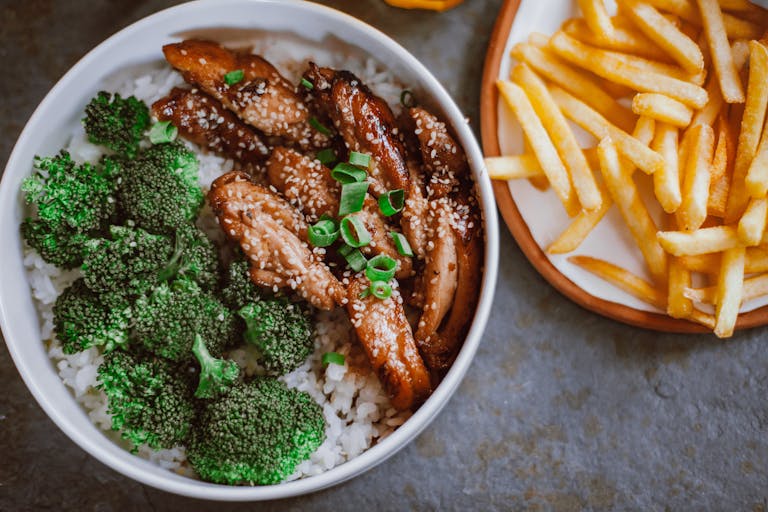 A tasty chicken, broccoli, and rice bowl garnished with sesame seeds, paired with crispy fries.