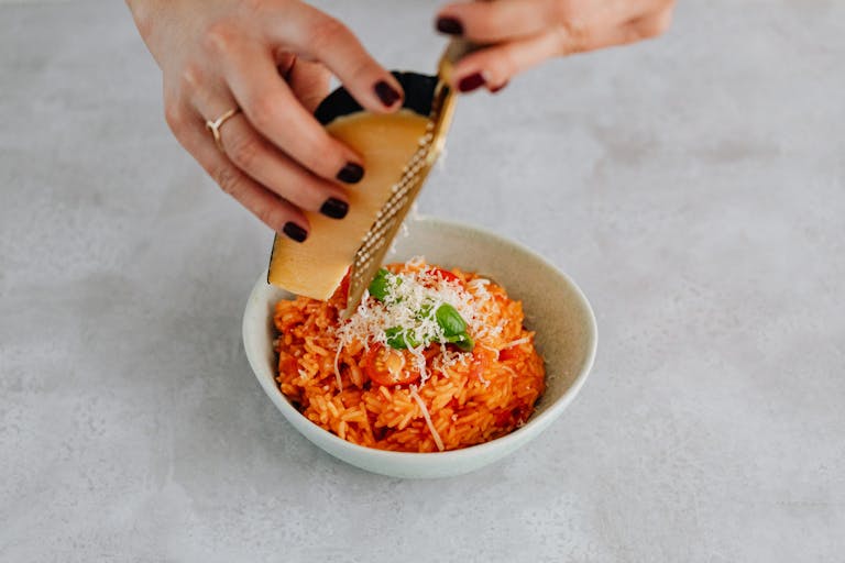 Close-up of hands grating parmesan cheese over a delicious bowl of tomato risotto.